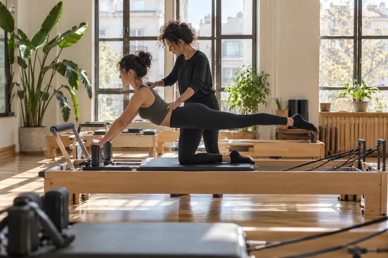 Aydınlık bir pilates stüdyosunda eğitmen, reformer aletinde çalışan öğrencisine yardım ediyor.